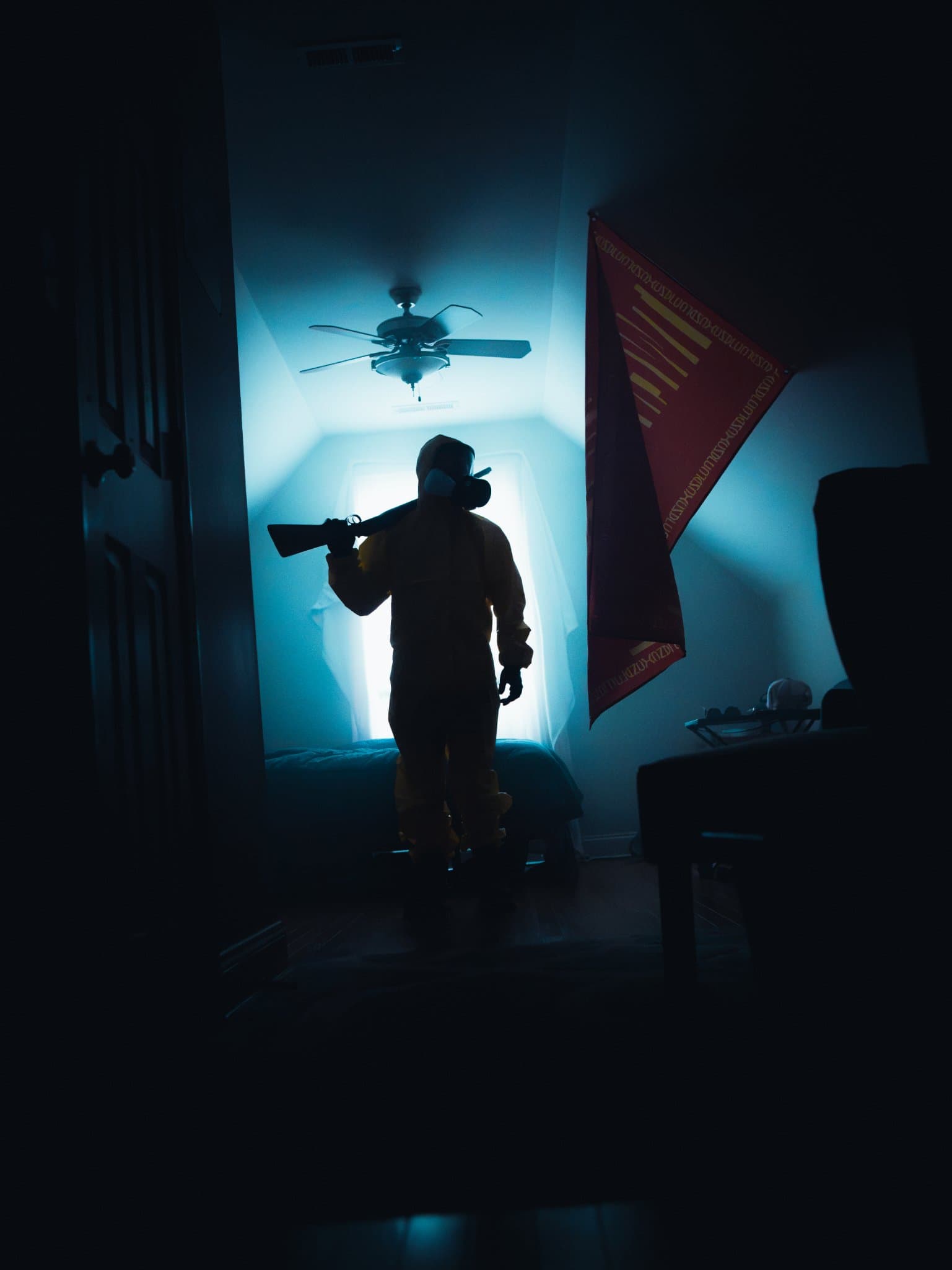 Dark full-body silhouette of a hazmat-suited figure with a rifle over one shoulder, framed by a doorway, a red flag hanging beside them — human-made reference photo on Slow Sync
